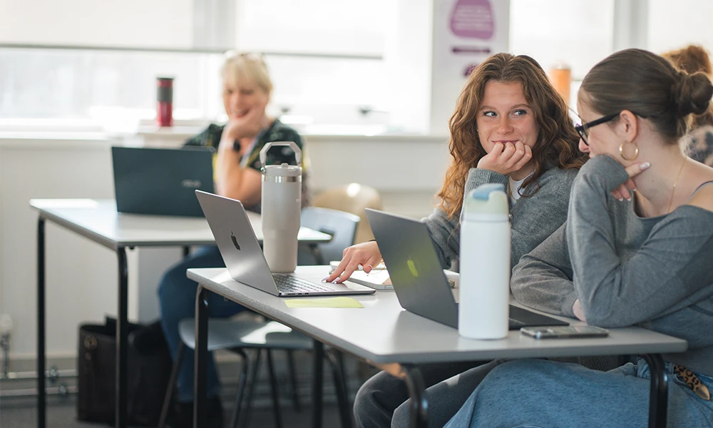 Two women sat in a classroom table with laptops infront of them