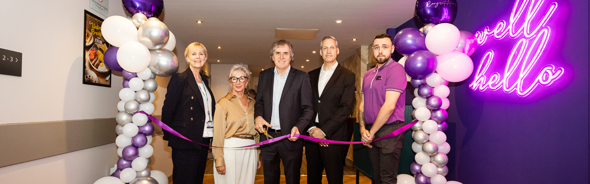 Five people Standing in front of a premire inn reception with a purple ribbon being cut.