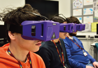 Three Computing And IT Students Wearing Virtual Reality Headsets In Classroom