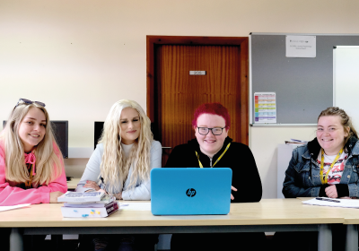 Four Wirral Met Social Studies students sat on a long table working on a blue laptop
