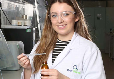 Female Wirral Met Science student smiling and standing in a lab wearing a white lab coat and safety glasses