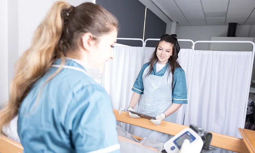 Two health and social care students practicing treatments on a maniken lying on a bed inside a classroom on Twelve Quays