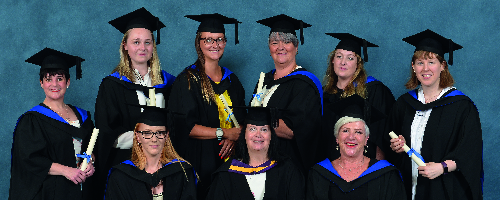 A group of Higher Education graduates wearing black gowns and caps and are holding scrolls, posing in front of a blue background